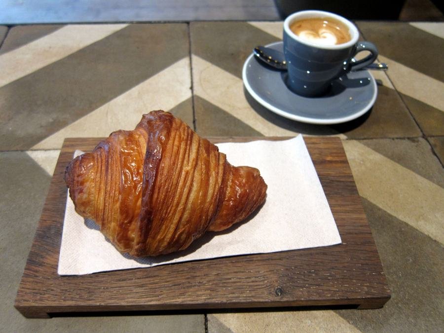 A glossy, flaky croissant on a wooden board with a cappuccino